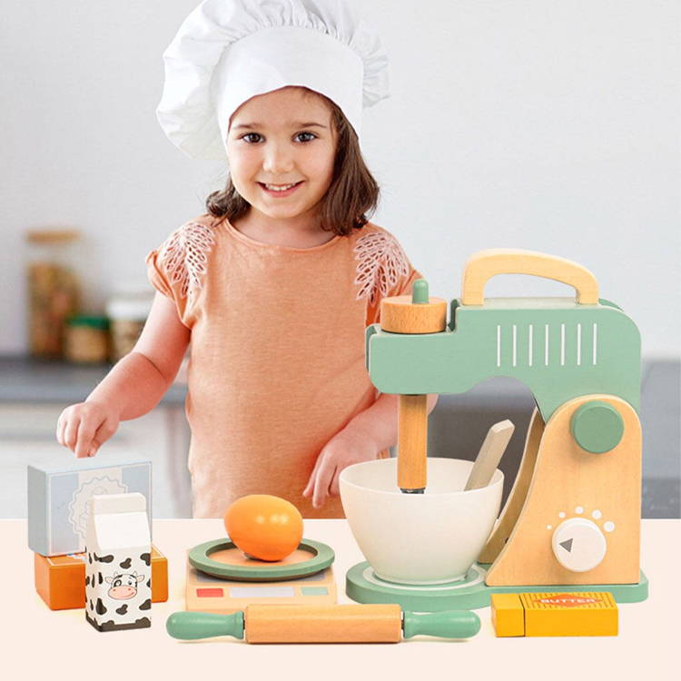 Toddler with a chefs hat next to the pretend play wooden mixer set.