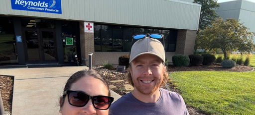 Two smiling individuals take a selfie outside Reynolds Consumer Products, holding boxes of food donations on a sunny day.