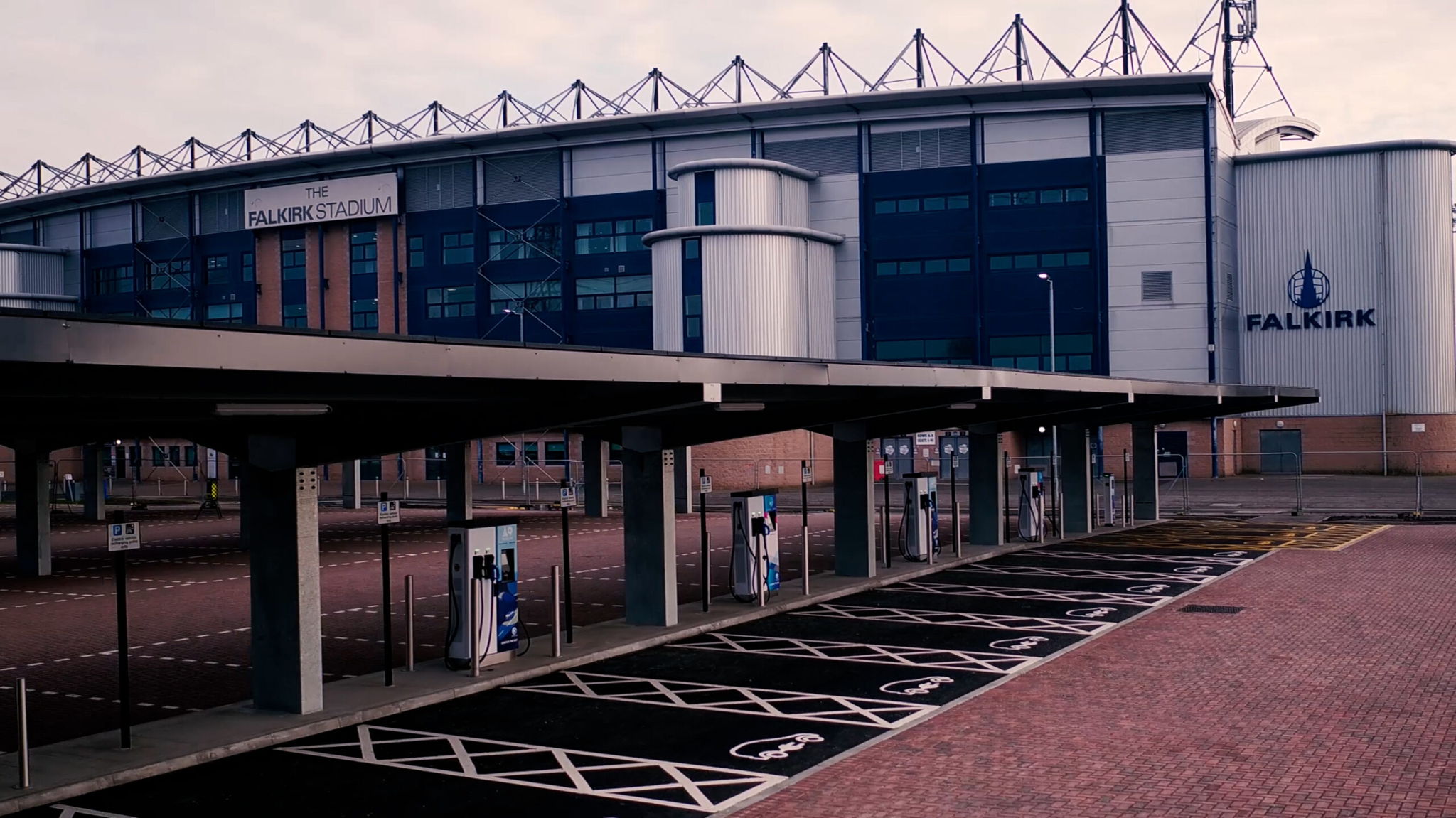 The image shows The Falkirk Stadium with a focus on its exterior. In the foreground, there is an empty charging station for electric vehicles with designated parking spots marked by white lines and symbols. The sky appears overcast.