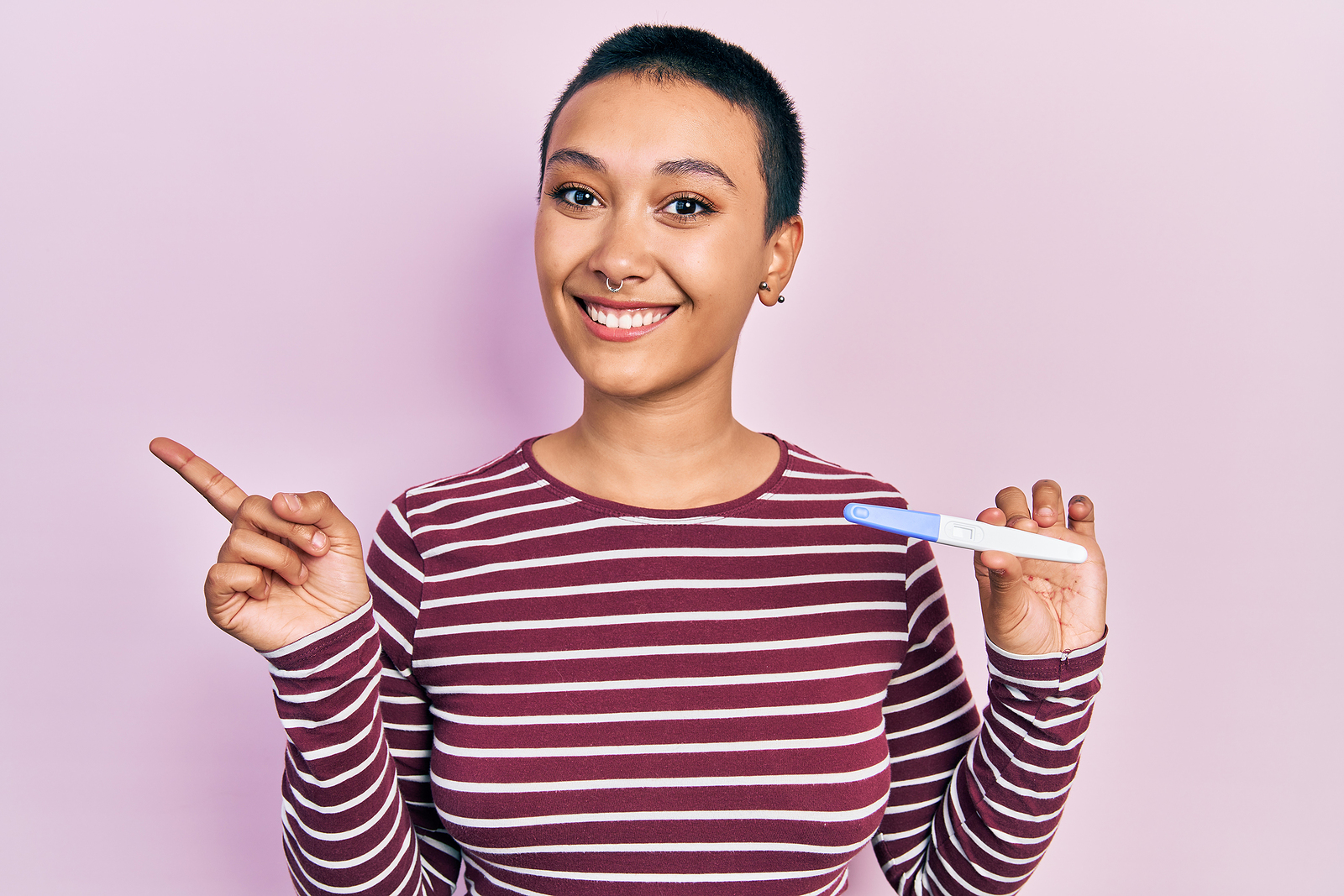 Beautiful hispanic woman with short hair holding pregnancy test result smiling happy pointing with hand and finger to the side