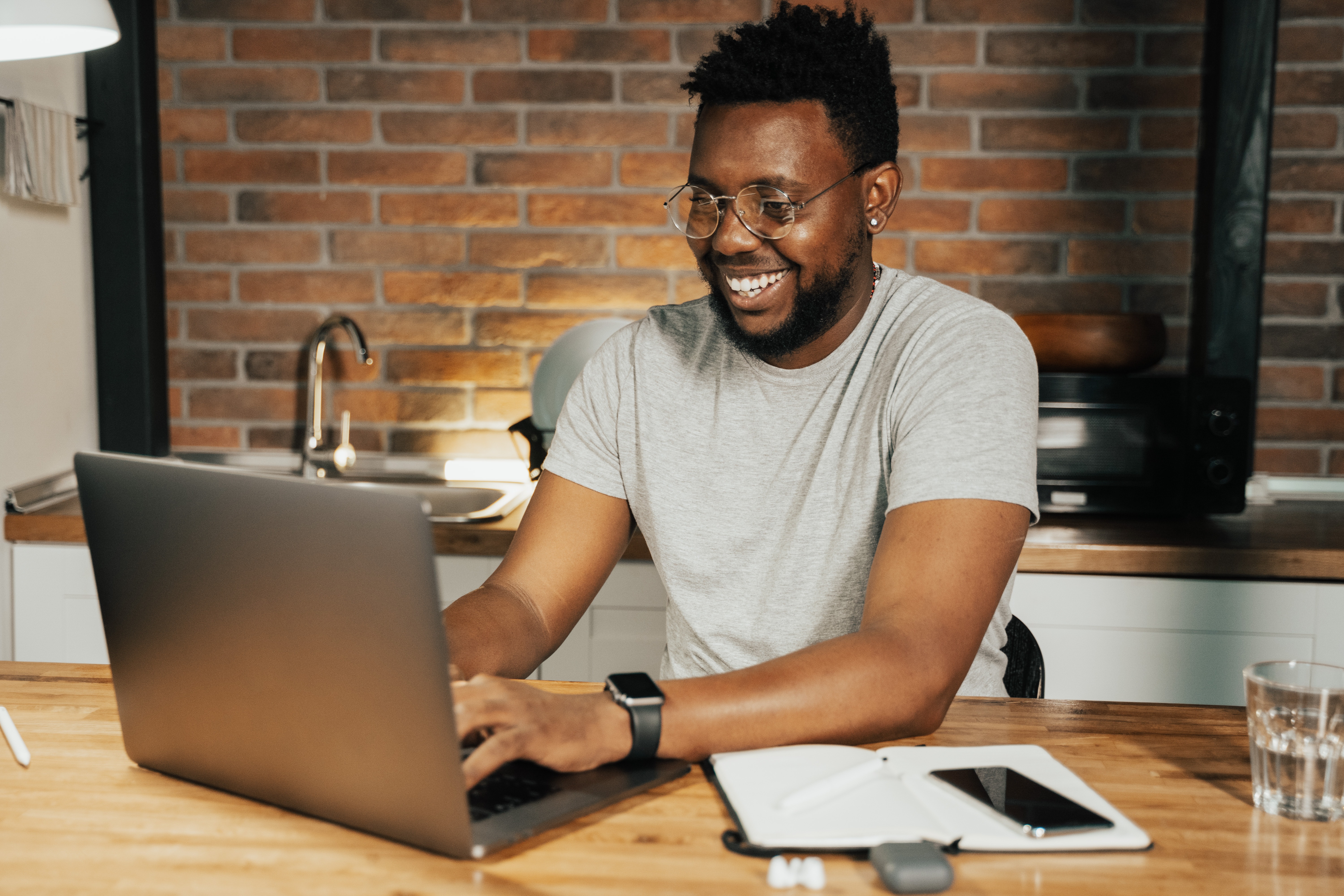 An attractive black man wearing glasses smilies while on his laptop on his kitchen counter.