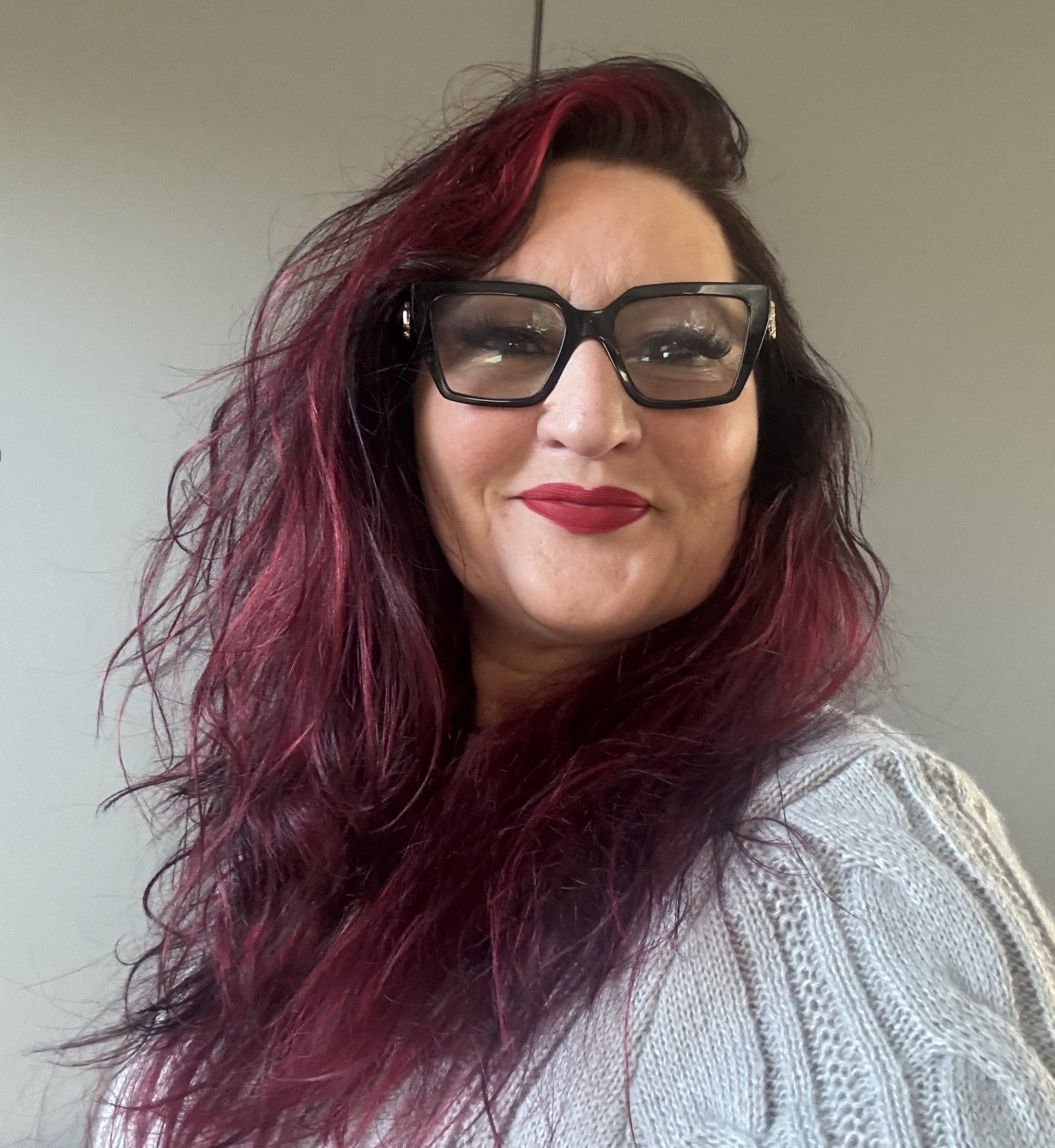 A woman with long, wavy dark hair with red highlights wears black glasses and a light gray knit sweater, smiling at the camera against a neutral background.