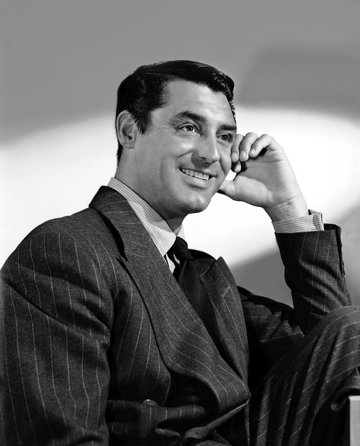 Black and white image of Cary Grant sitting on a chair smiling with a hand on his cheek.