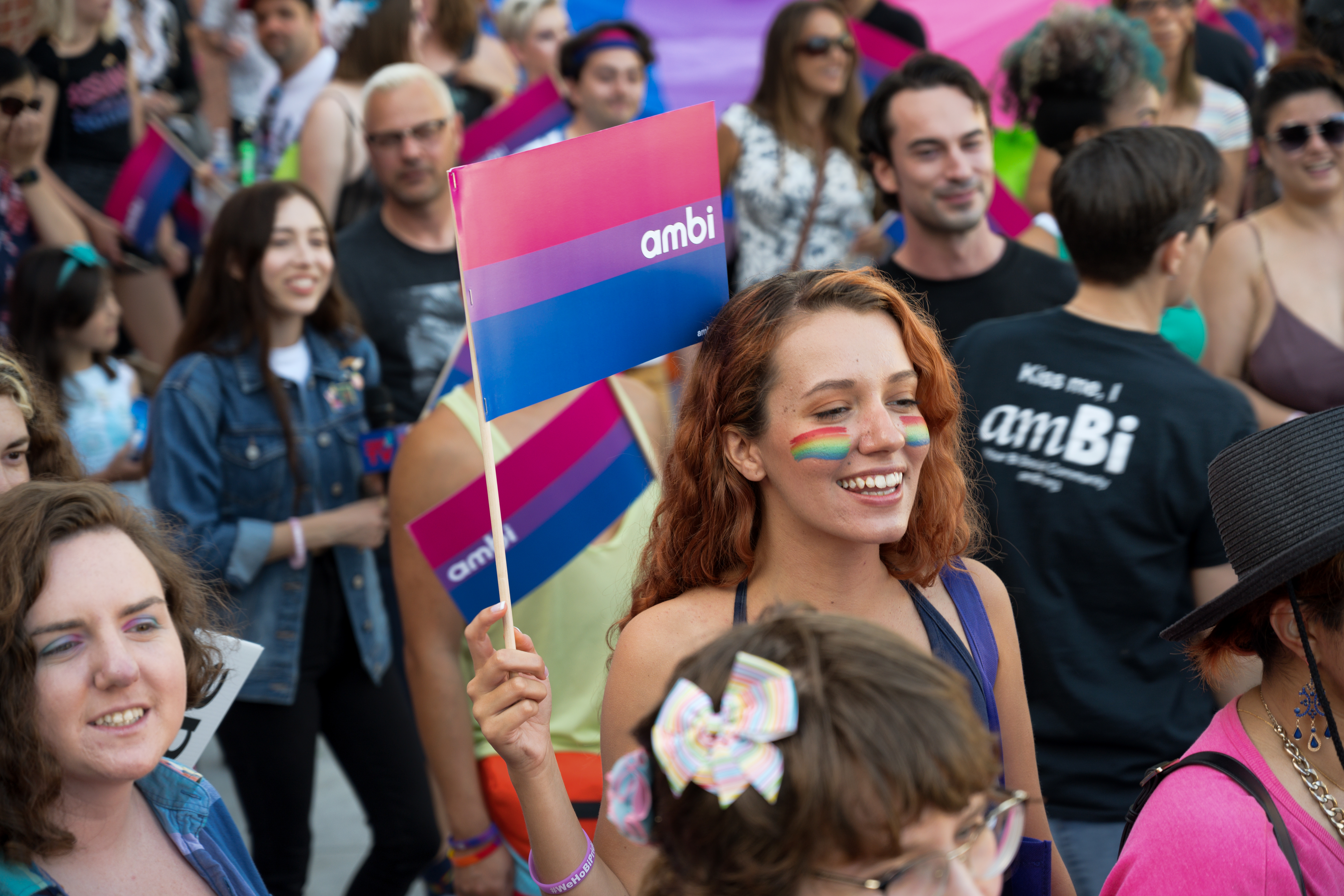 During Pride, a large group marches and a woman holds a bi flag above her head smiling.