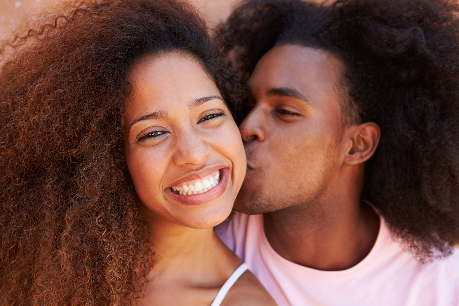 A dark skinned couple with large curly hair, the girl is smiling at the camera and the man is kissing her cheek.