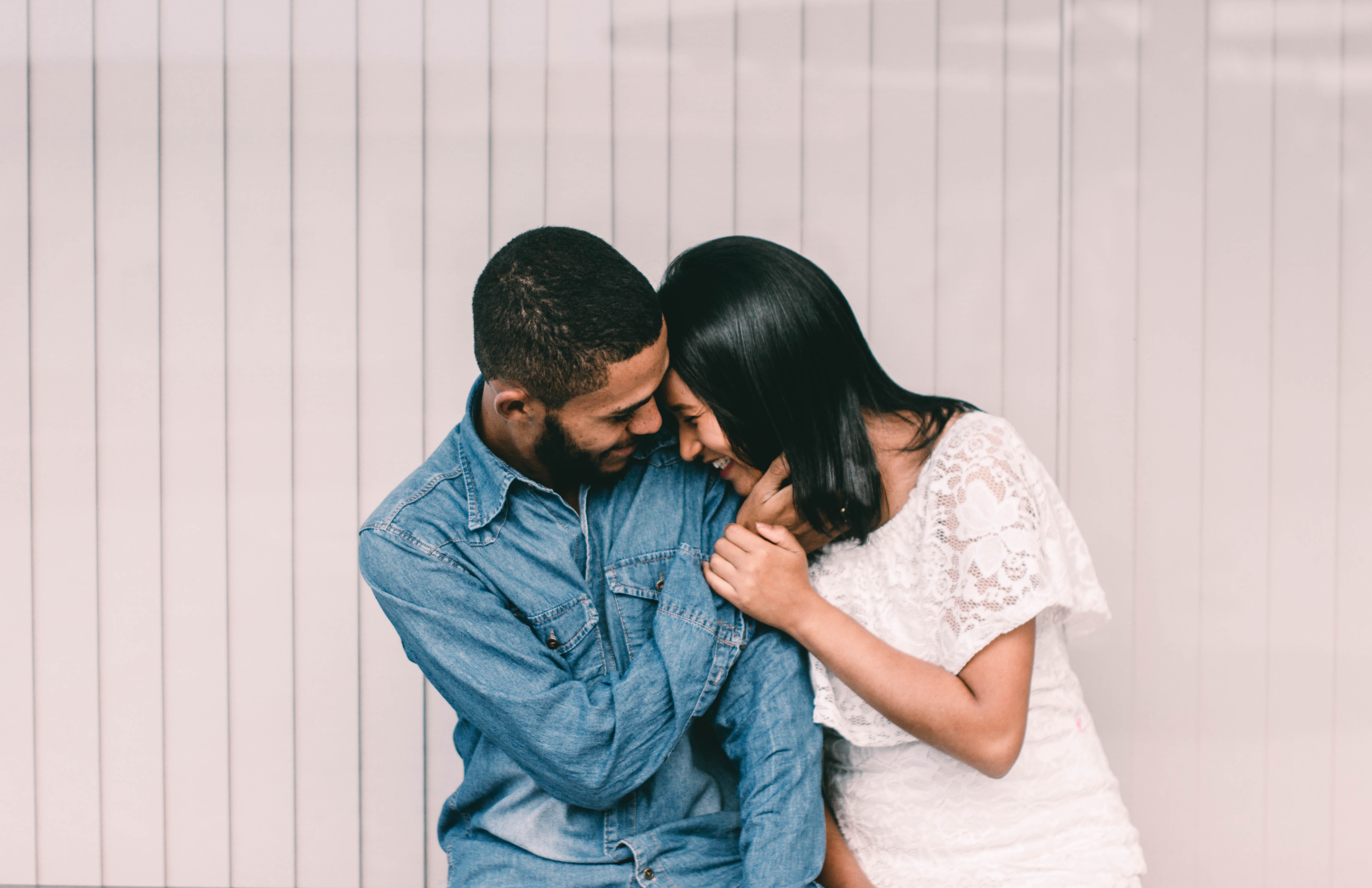 A multi ethnic couple laughs together and leans in closely while touching eachothers' faces.