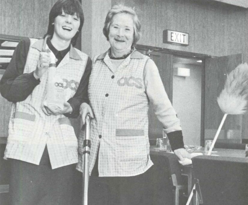 Two women in checkered uniforms smiling, one holding a spray bottle and a vacuum, the other leaning on a broom. A sign reading EXIT is visible in the background. They appear to be in a cleaning setting.