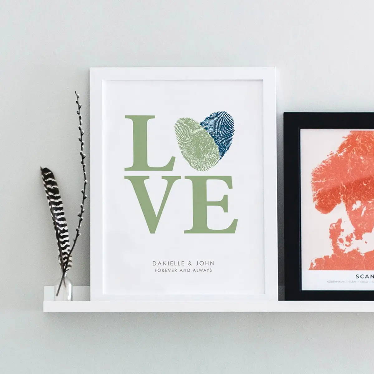 Two framed prints on a white shelf: one says "LOVE" with fingerprint heart, another shows a red map of Scandinavia. A feather and a small plant decorate the shelf.