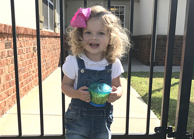 Little toddler girl smiles as she stands outside the school gates on her first day