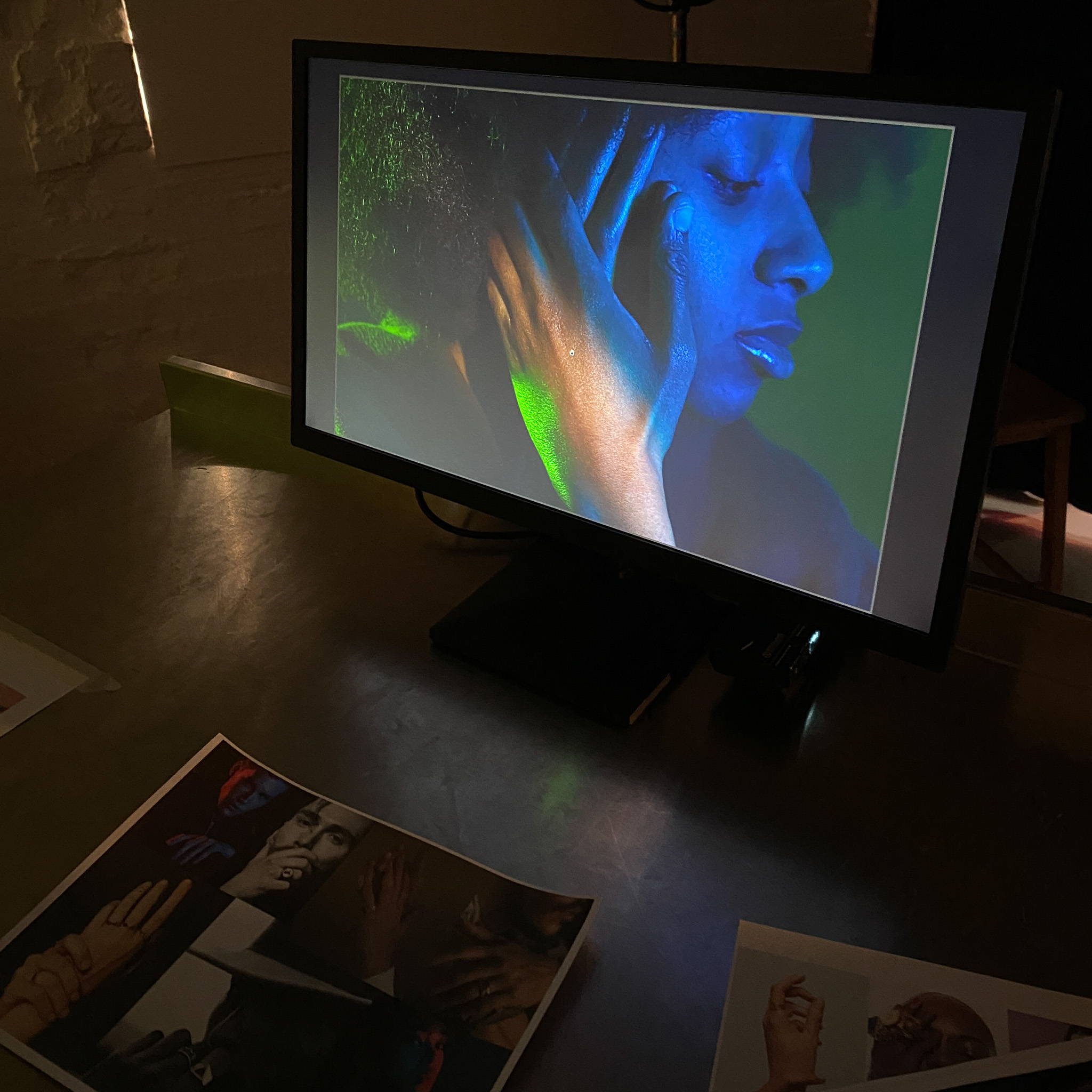 A monitor screen sits on top of a table. In the foreground, two pieces of paper are also on the table. These feature a collage of moody images for inspiration. On the monitor, model Rhianna is bathed in green and blue lighting and holding their hands up to the right side of their face which is turned to the side.