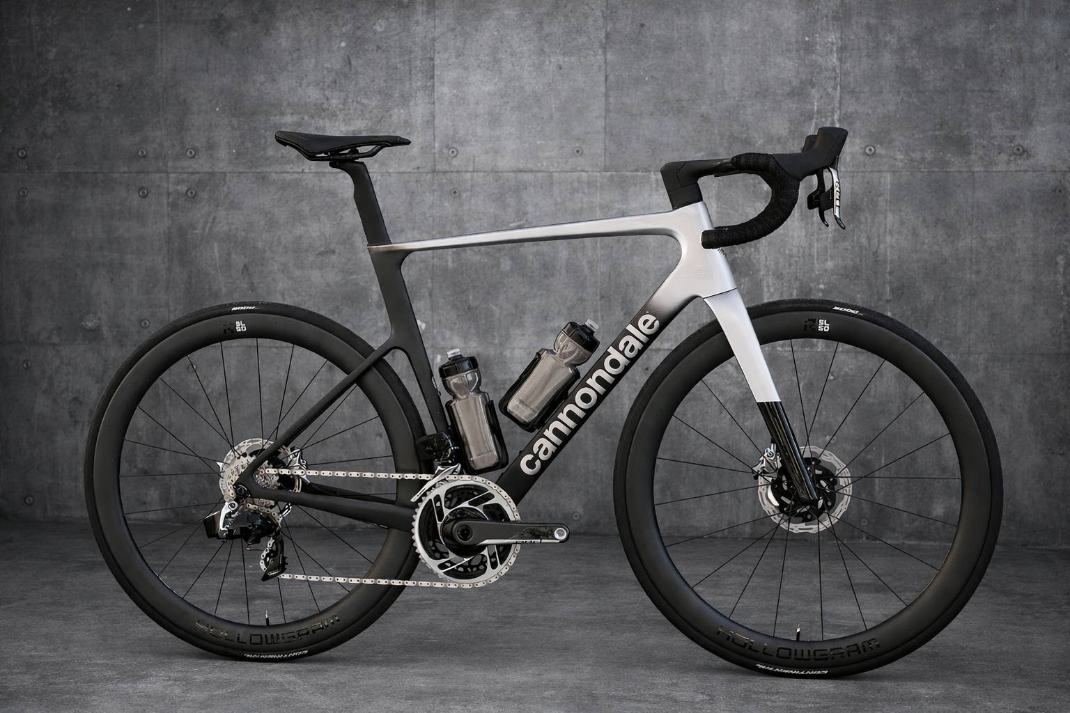 Cannondale SuperSix EVO racefiets in zwart-zilver zijaanzicht tegen een industrieel grijze betonnen achtergrond, met diepe carbon velgen, schijfremmen en bidons in het frame.