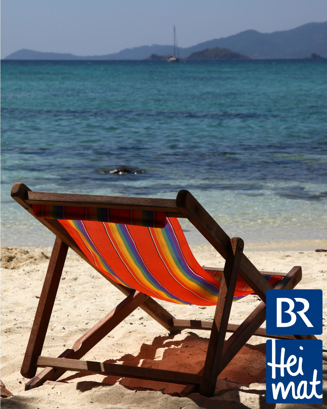 Ein bunter Liegestuhl steht am Strand. Im Hintergrund ist das Meer zu sehen und die Sonne scheint. 