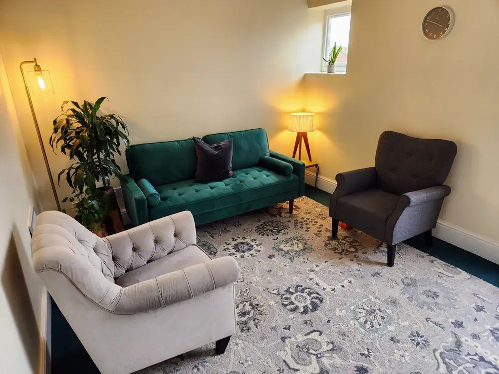 Photo of the therapy room in which James Morris Psychotherapist in Poole consults with clients. The room contains a green sofa flanked by single seater armchairs with lamps and a pot plant to the side of the sofa.