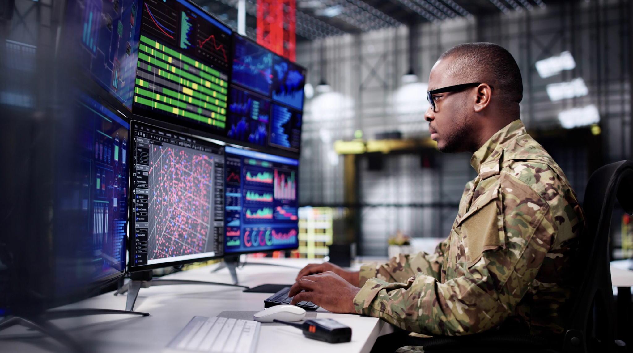 A person in military uniform works at a desk with multiple large monitors displaying data, graphs, and maps in a modern command center or control room environment.