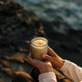 A person - can only see their knees and hands - holding a lit St Eval candle in glass, sitting on a rock on a beach - the sea can be seen blurred.