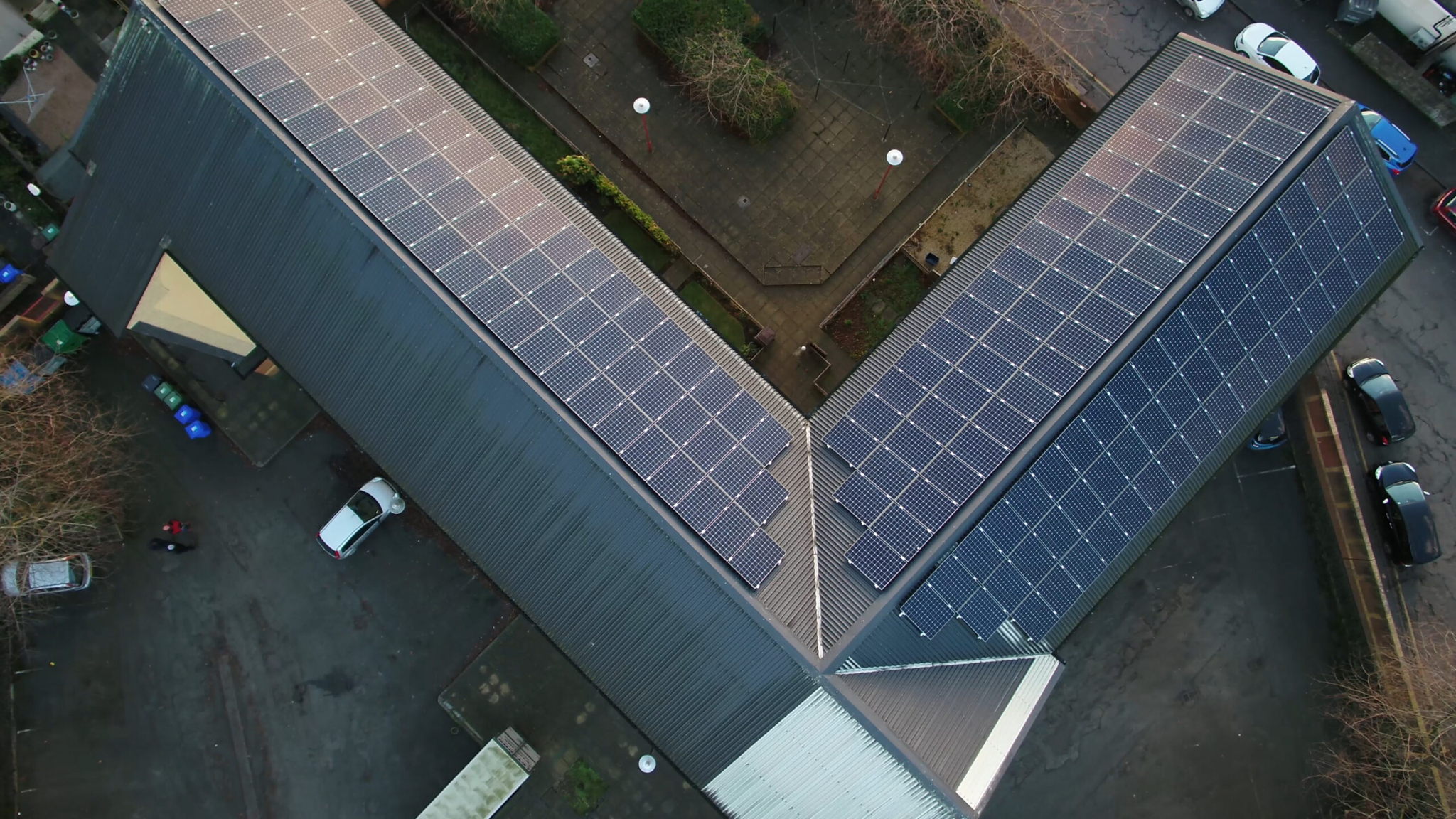 Aerial view of a building with a V-shaped roof covered in solar panels, surrounded by pavement, greenery, and a parked white car.