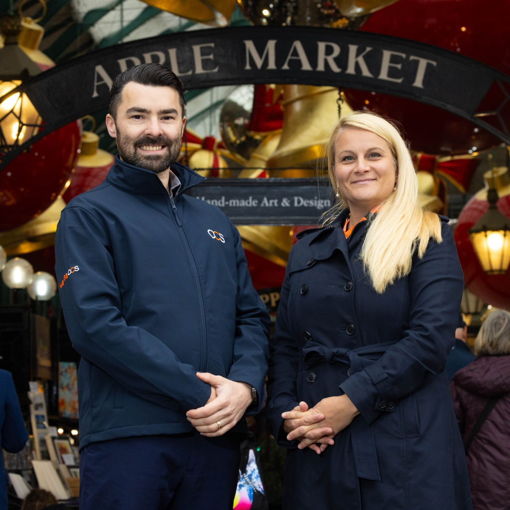 Two people stand smiling in front of the Apple Market entrance, wearing navy blue jackets. The market sign and festive decorations are visible in the background.