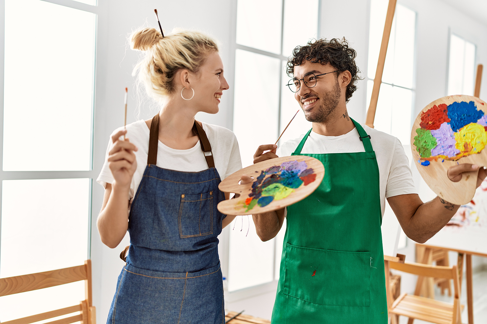 An attractive man and woman laugh as they hold paint brushes in their hands. They are wearing aprons.