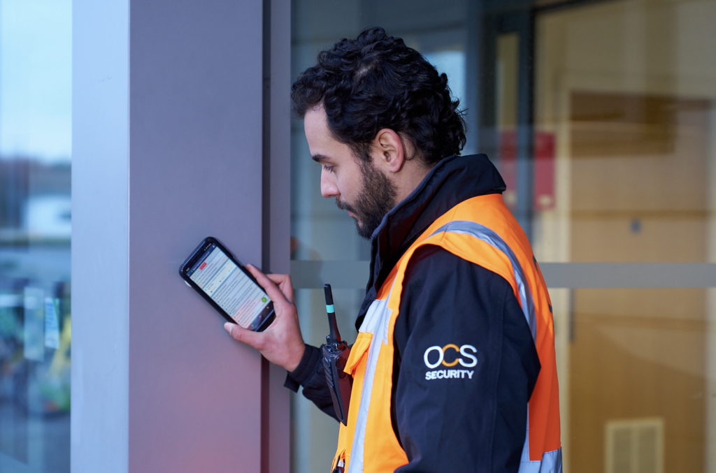 A security guard wearing an orange high-visibility jacket and holding a walkie-talkie checks a smartphone while standing next to a glass door. The jacket is labeled “OCS Security.”.