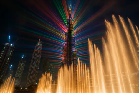 Dubai's Burj Khalifa building illuminated by a vibrant rainbow light display and a powerful yellow water fountain show at night.