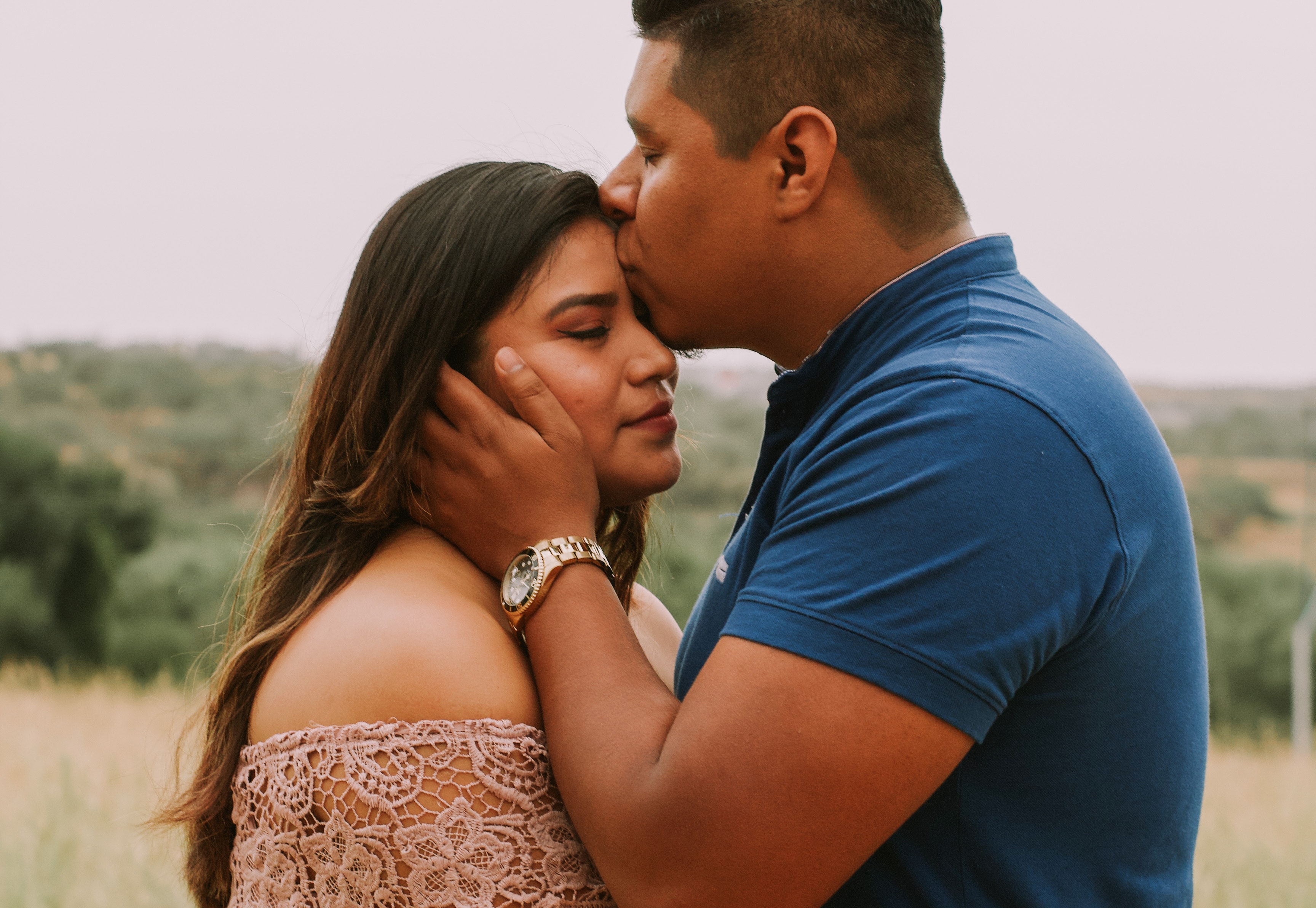 A couple standing in a meadow. The man is holding his girlfriend's head and giving head a kiss in the forehead.