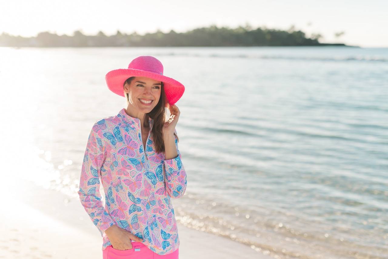 A woman wears a butterfly patterned pink and blue cover-up, pink shorts and a matching pink hat walking along the beach.