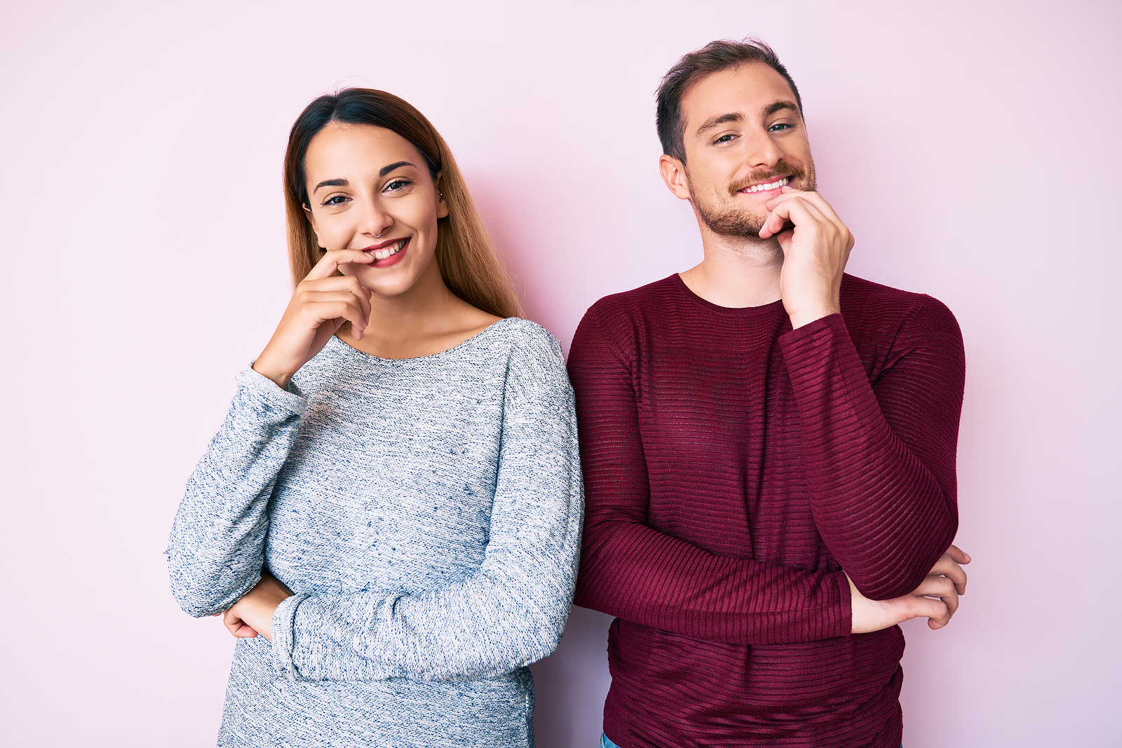 An attractive man and woman smile curiously while holding a hand to their chins and cross their arm.