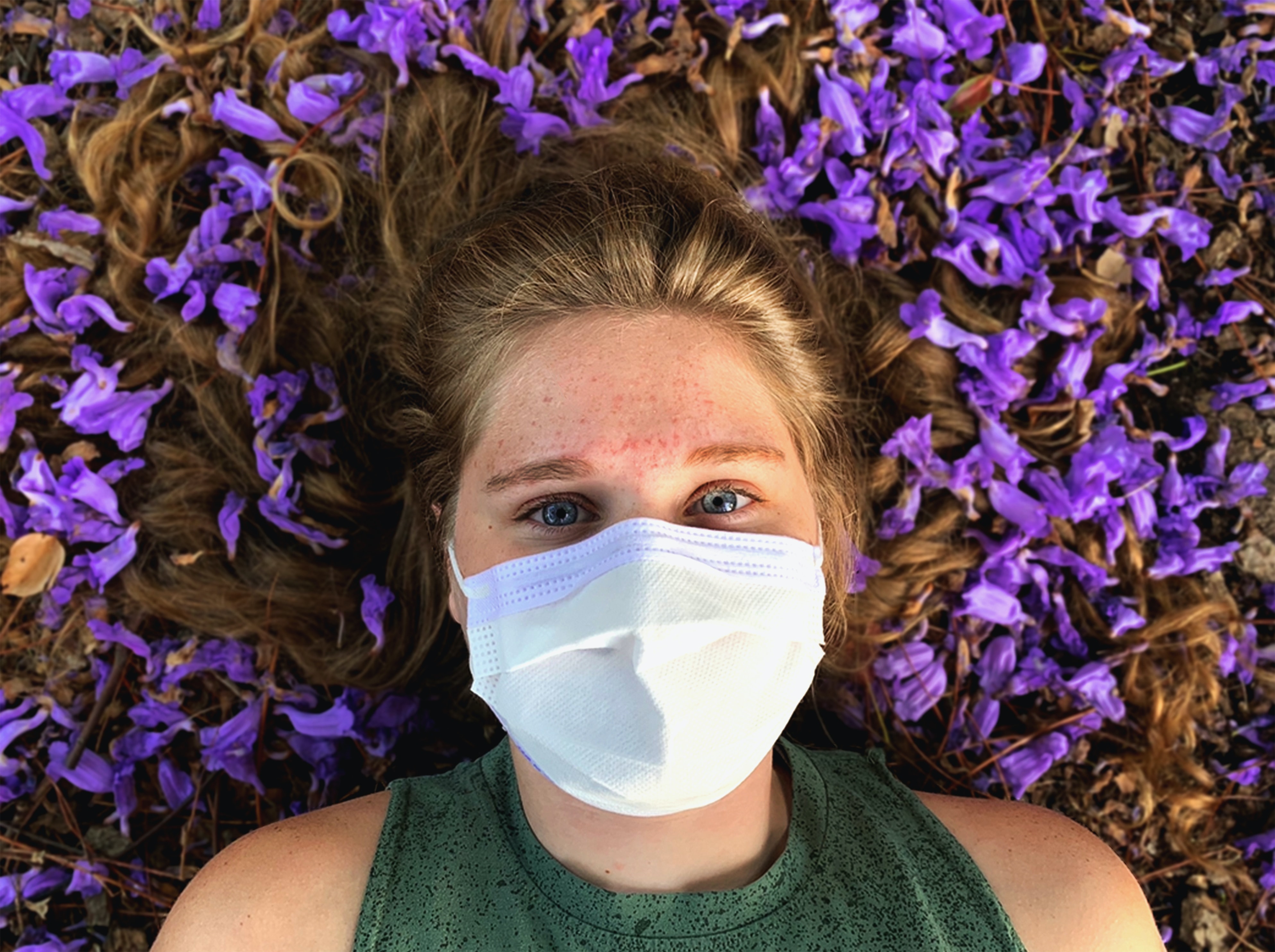 A blond haired woman wearing a face mask lays near flowers.