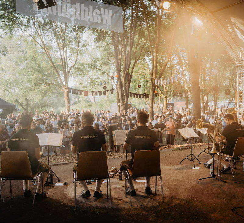 Auf einer Festival-Bühne im Freien sitzen 5 Blasmusiker. Vor ihnen steht Publikum. 