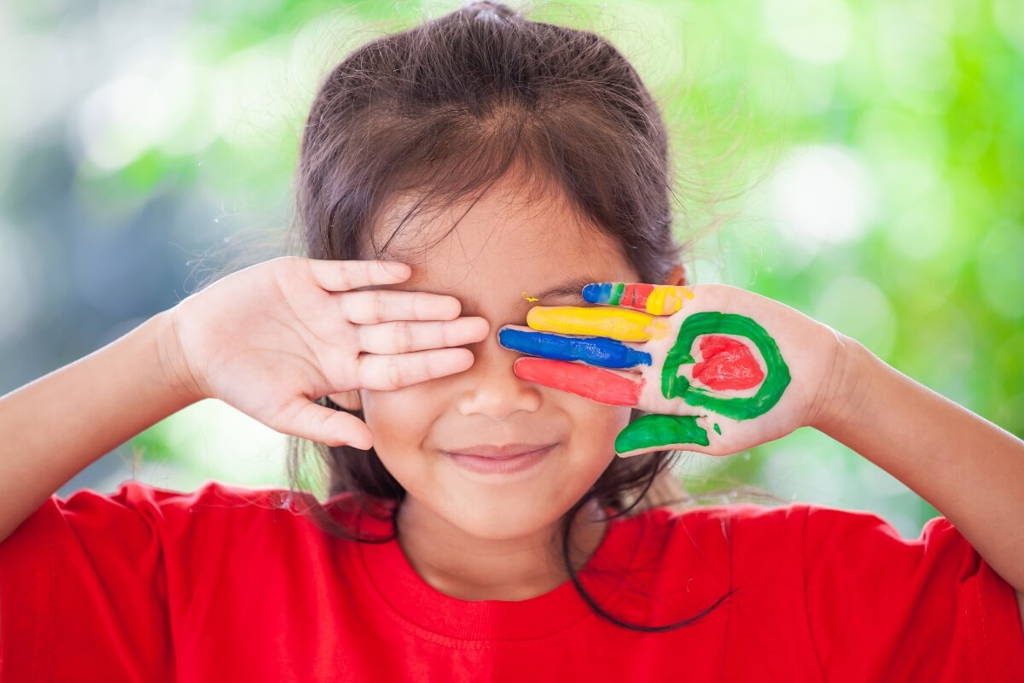 Une jeune fille souriante, vêtue d'un t-shirt rouge, se couvre les yeux avec malice, une main peinte de rayures et de formes colorées, se détache sur un fond extérieur vert flou.