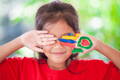 Smiling girl in a red shirt playfully covering her eyes, one hand painted with colorful stripes and shapes, against a blurred green outdoor background.