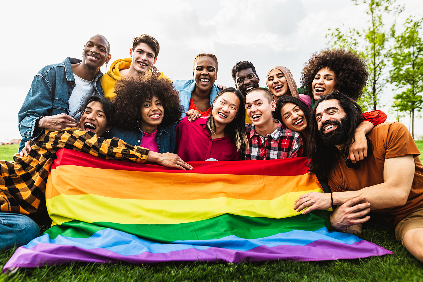A large group of friends all smile and embrace eachother as they sit on the grass together holding a rainbow flag.