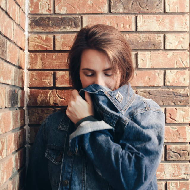 Brunette girl against a red brick wall wearing a jean jacket with enamel pins