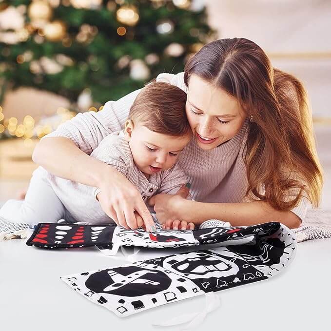 Une jeune femme et un nouveau-né explorent joyeusement ensemble un livre de contraste pour nouveau-né InvenToy Montessori dans une pièce chaleureuse décorée d'un sapin de Noël, transmettant une atmosphère chaleureuse et festive optimisée pour la stimulation visuelle et le développement de bébé.