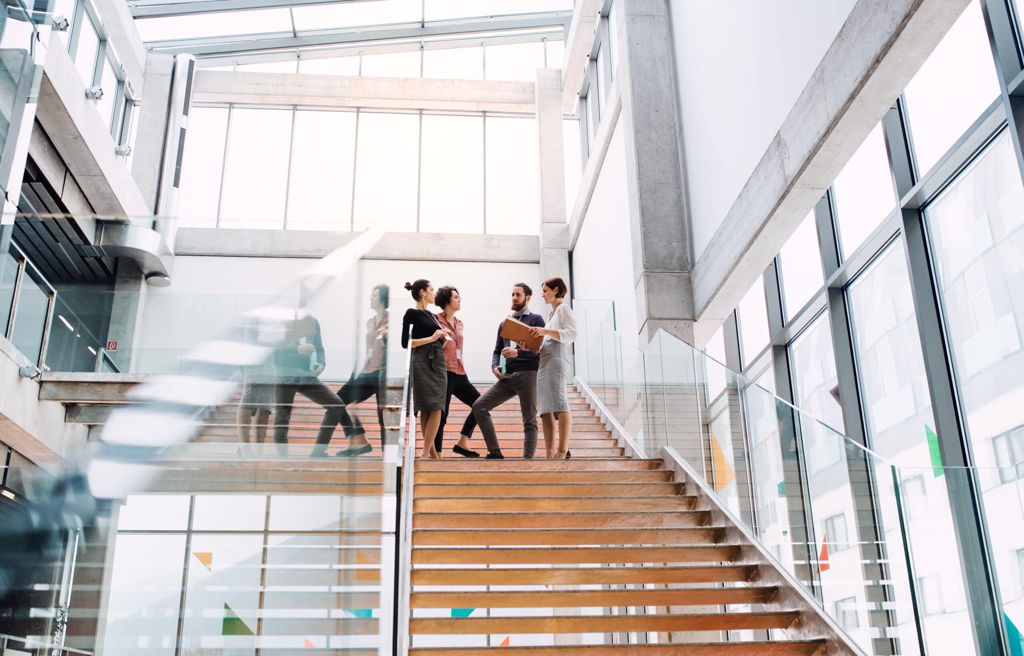 Four people stand on a modern stairway in a bright office building, engaged in conversation. The space features large windows, glass railings, and an airy, open design.