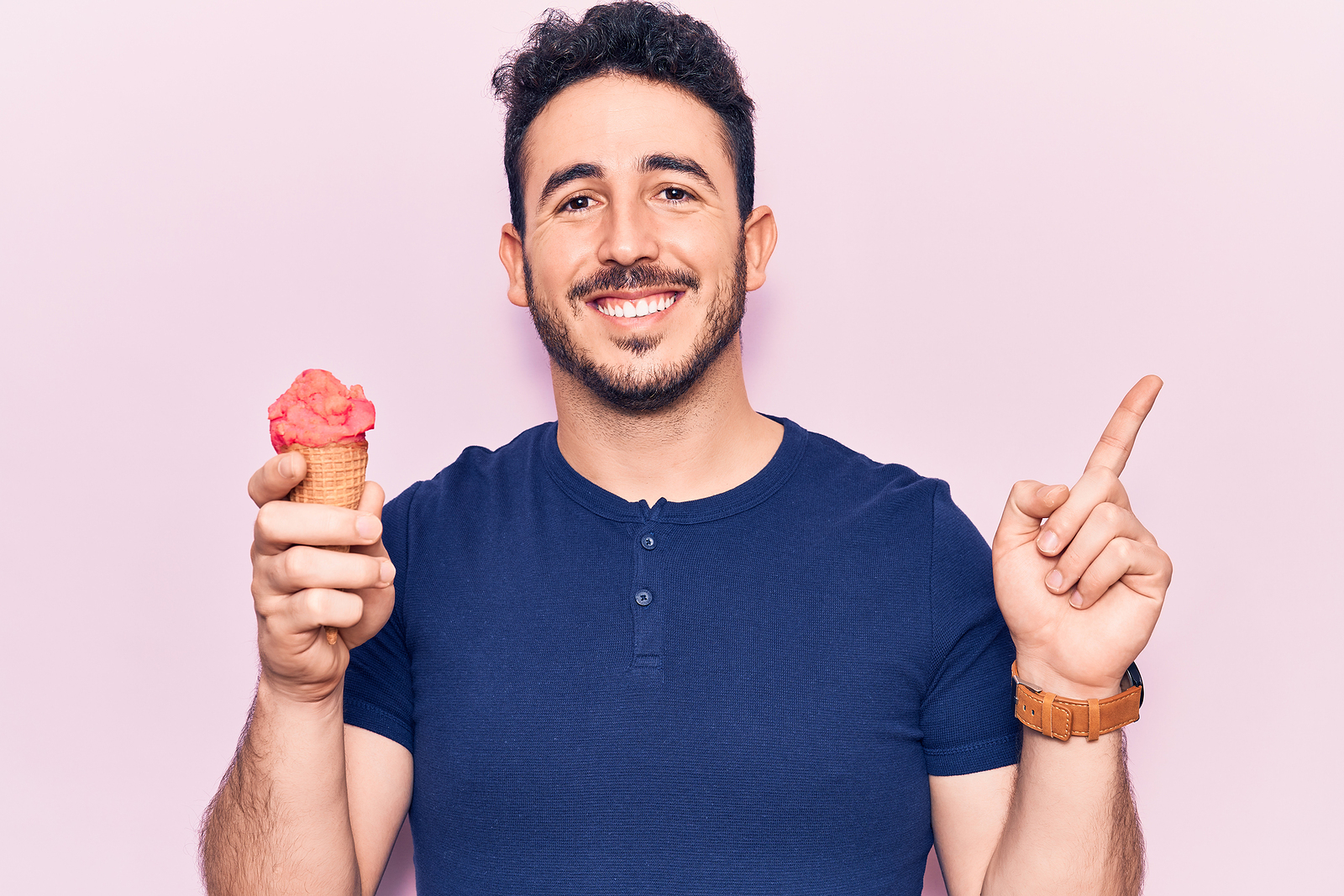 A young attractive man smiles while holding an ice cream cone and pointing to the other direction with his other hand.