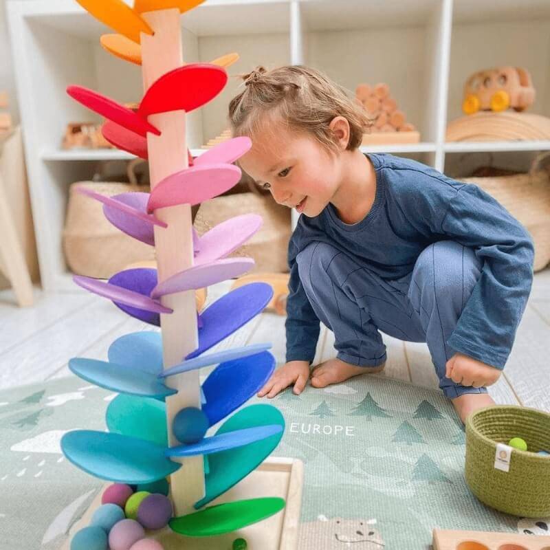 Un jeune enfant vêtu d'une tenue bleue sourit en jouant avec un arbre arc-en-ciel Montessori InvenToy dans une salle de jeux bien organisée.