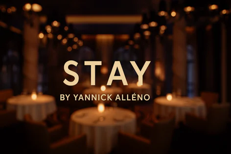 Text overlay reads "STAY BY YANNICK ALLÉNO" against a blurred background of a fine dining restaurant with tables and warm lighting.