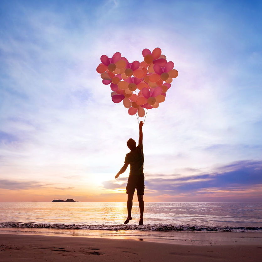 A silhouette of a man on a beach when the sun is low, being lifted by a large amount of heart balloons arranged into a heart.