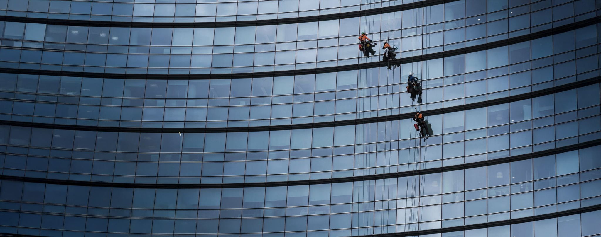 Four window washers wearing safety harnesses clean the glass windows of a tall, curved modern building, suspended by ropes several stories high against reflective blue-tinted glass panels.