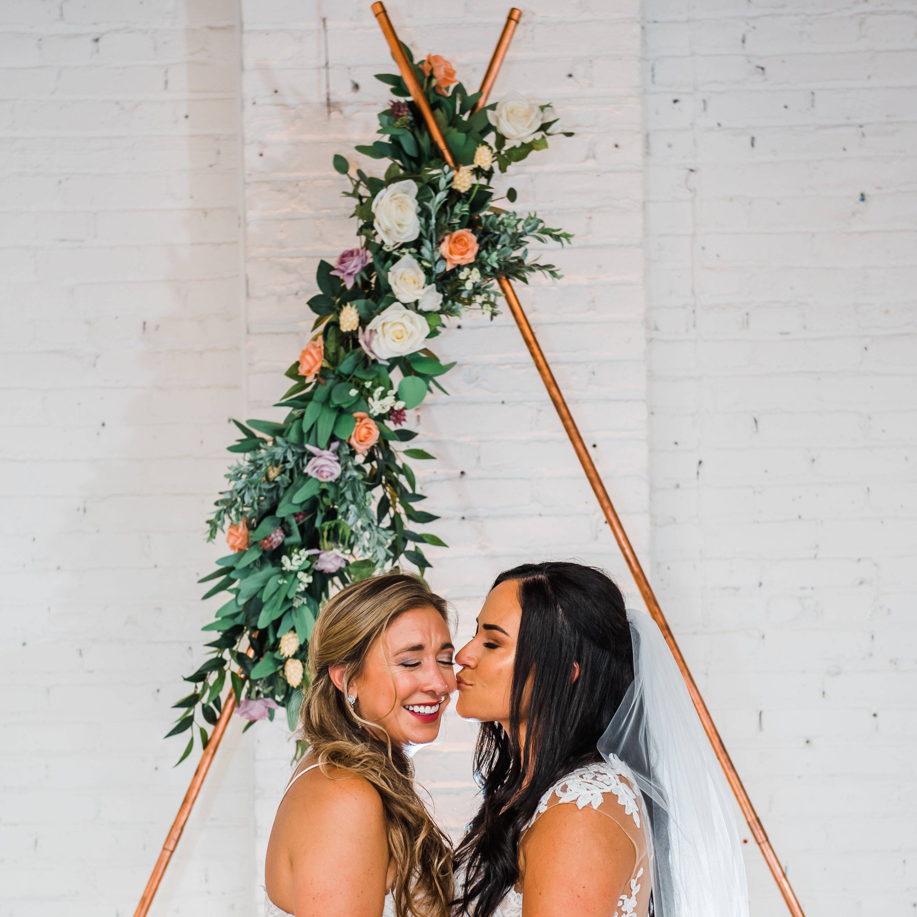 Two brides kiss under our triangular copper arch with purple and peach florals decorating it 