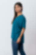A woman wearing a teal short-sleeve top with lace shoulder details and blue jeans, posed in profile against a light grey background.