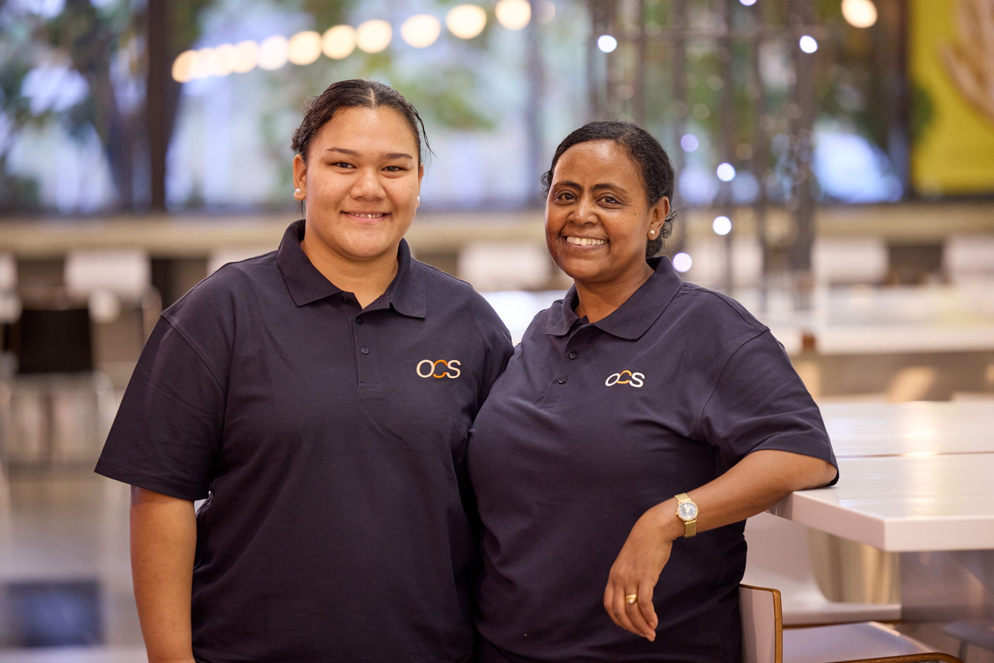 Two women wearing matching navy OCS polo shirts stand side by side indoors, smiling at the camera. The background is softly lit with blurred lights and greenery visible through windows.