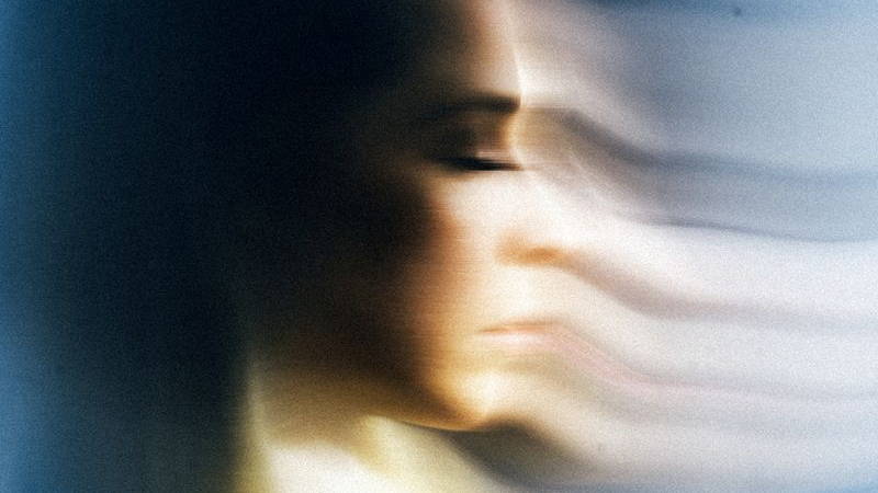 The woman's face is captured in profile with her eyes closed, her outline receding into a horizontally blurred, striped phantom image against a pale background. This conceptual portrait symbolizes psychological trauma, memory loss, dissociation, and the p