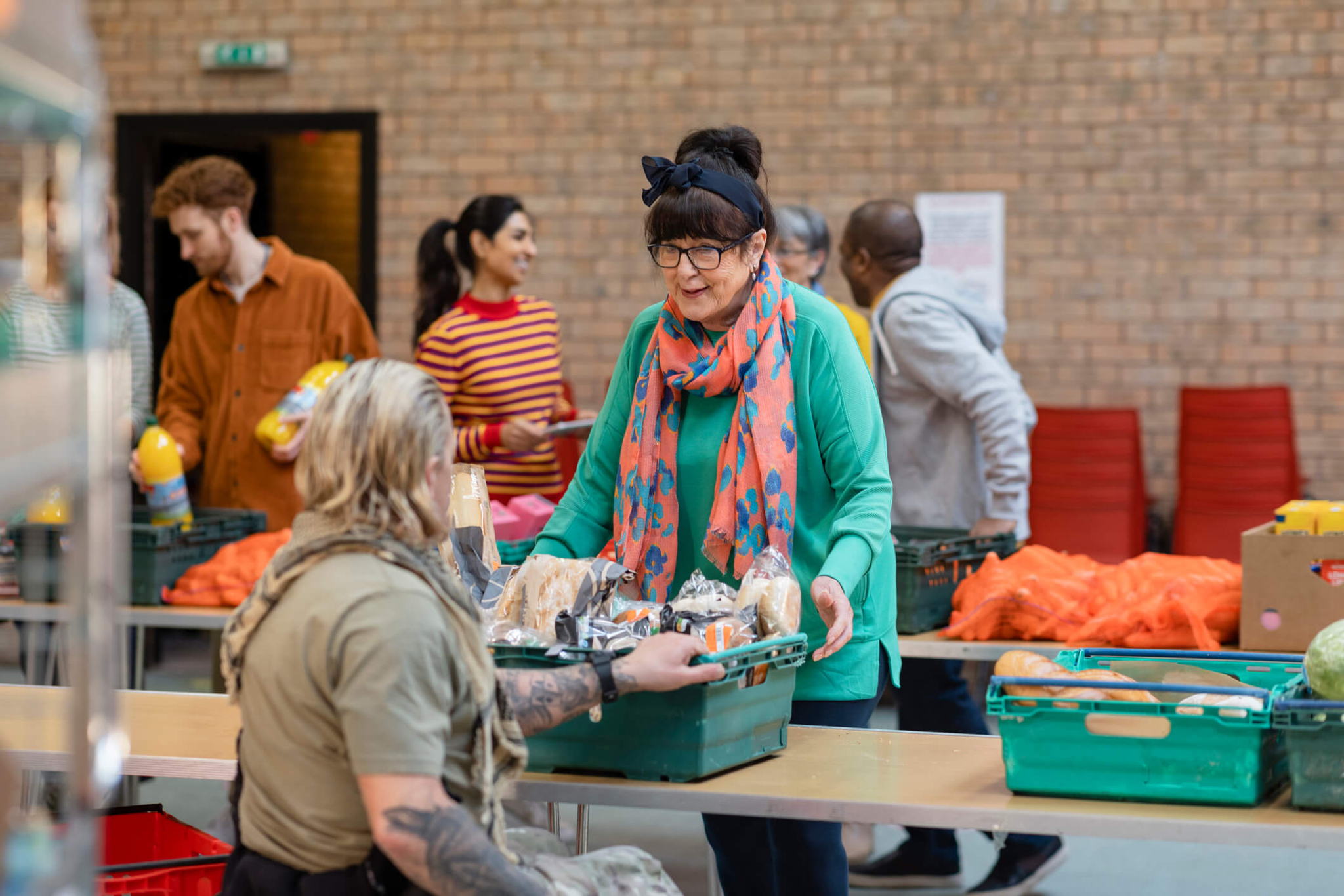 A woman with a colorful scarf and glasses hands a box of food to a seated person. Others are in the background organizing produce. The scene appears to be a community food distribution event in a hall with brick walls.