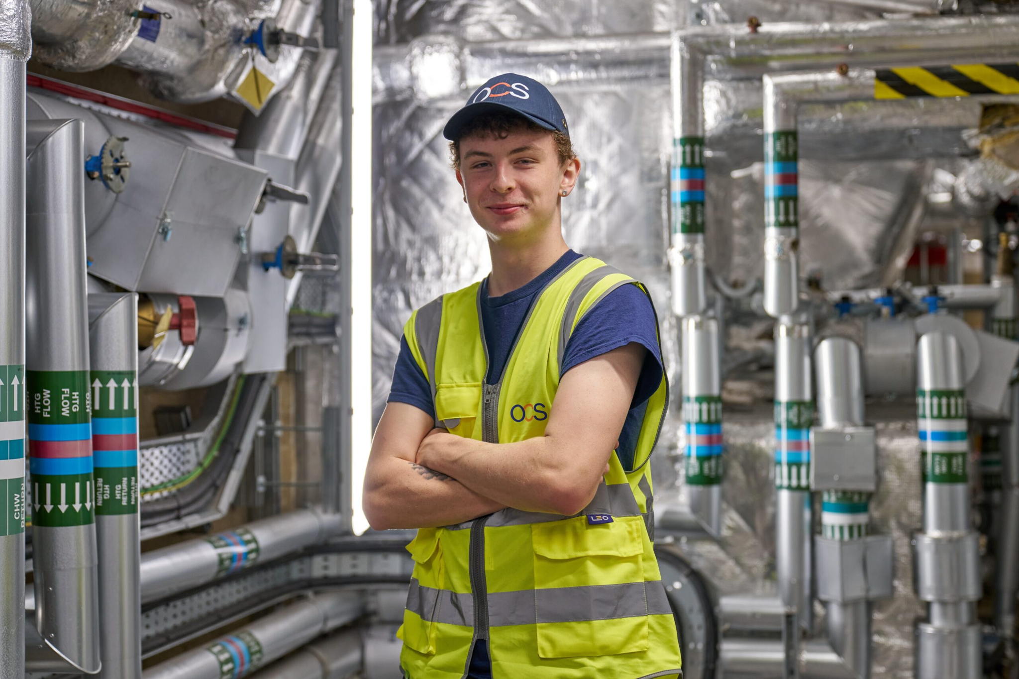 A young worker in a yellow safety vest and cap stands with arms crossed, smiling, in an industrial setting filled with metal pipes, ducts, and machinery.
