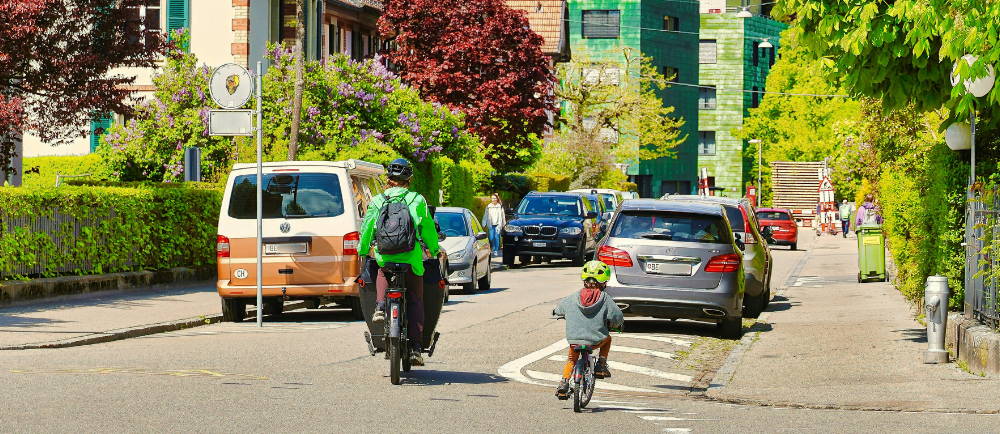a parent and child riding on a street lined with cars and bright trees