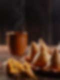 Plate of crispy, golden samosas arranged in a circle, with tamarind sauce being drizzled onto one samosa, and a clay teapot and steaming cup in the background, creating a warm and inviting scene.