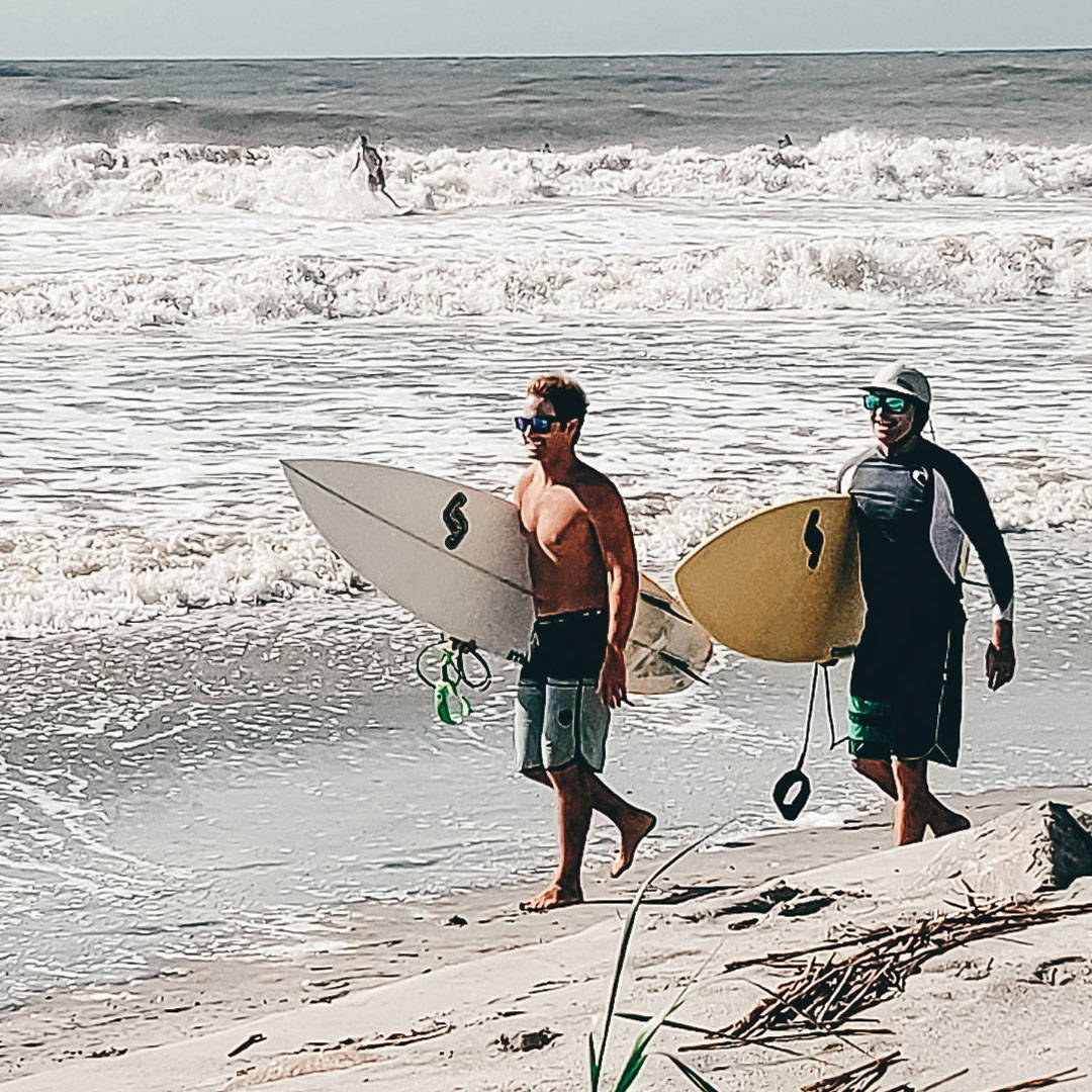 Two surfing buddies walk along the shore, wearing their Rheos sunglasses.