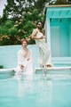 a man in a headscarf and caftan sits on the pool edge while a man in white sunglasses and a yellow midi shift dress perches on the handrail above for "poolside pride: a slim aarons-inspired photo campaign."